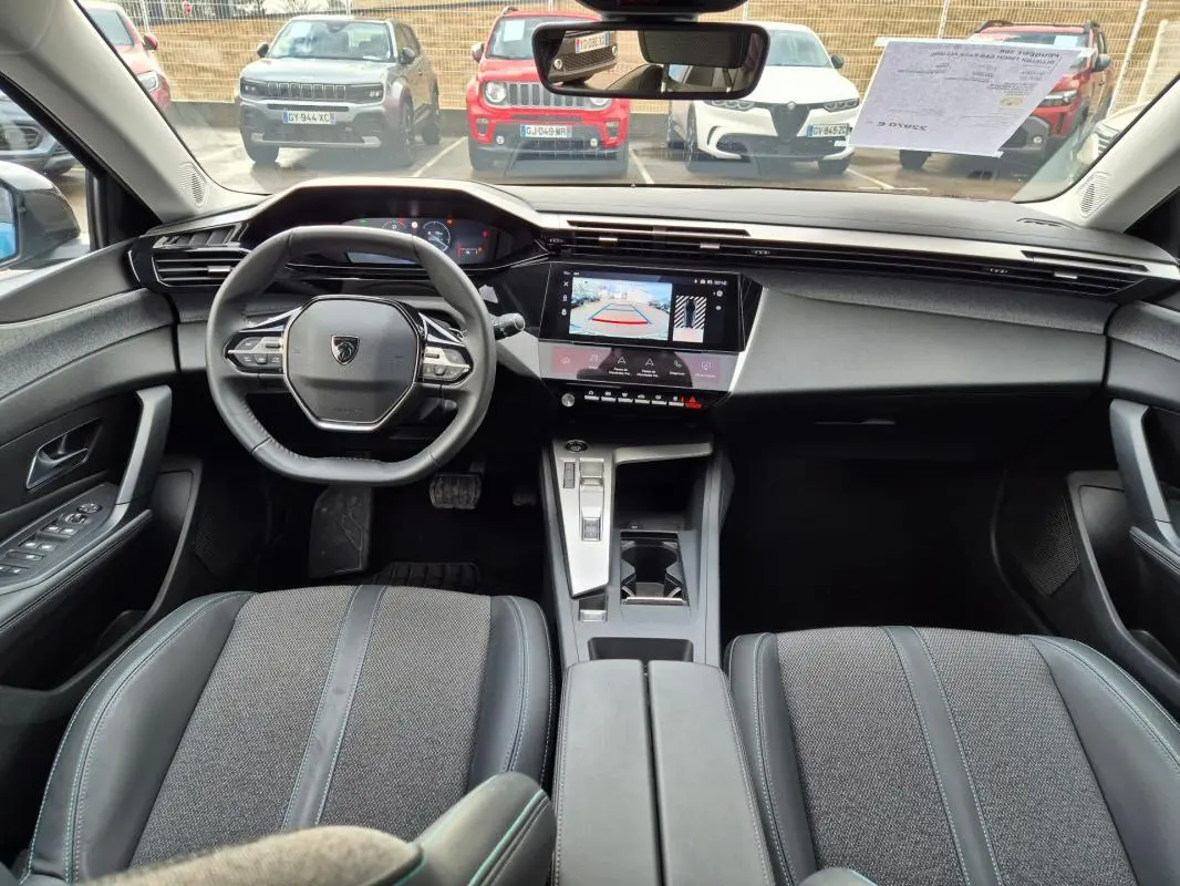 Vue intérieure avant du Peugeot 308 2024 bleu, montrant le volant compact, l’écran tactile avec caméra de recul et l’accoudoir central.