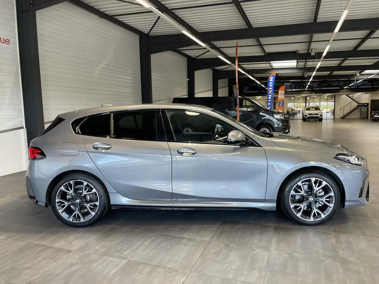 BMW Série 1 120d M Sport gris vue de profil côté gauche dans un showroom lumineux avec jantes sport.