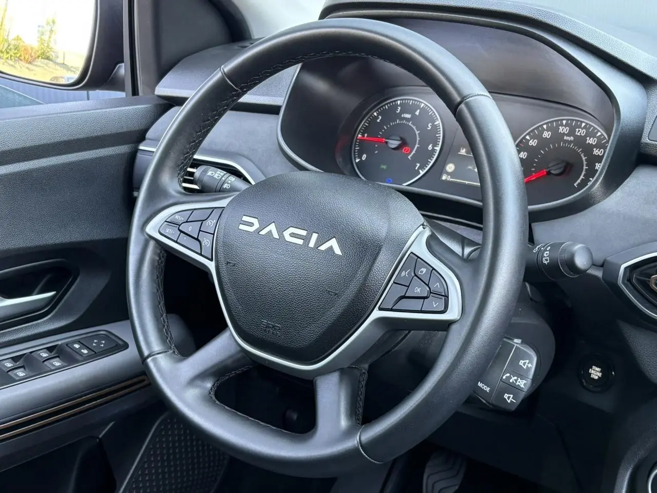 Vue intérieure centrée sur le volant noir Dacia avec commandes intégrées et tableau de bord analogique.