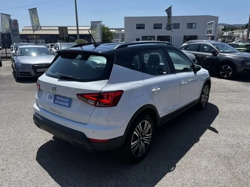SEAT Arona blanc 2024 en 3/4 arrière droit, toit noir contrastant et jantes alliage visibles sur parking.