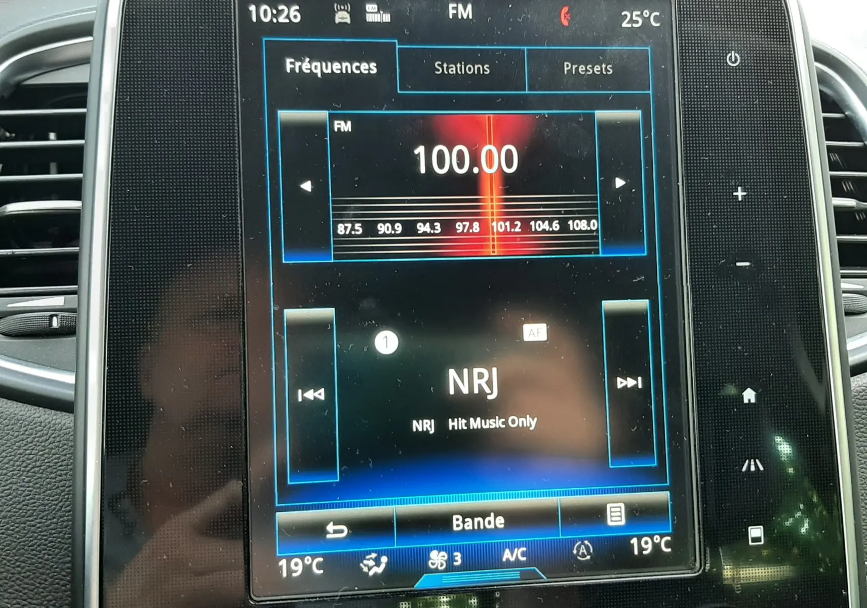 Écran central tactile du Renault Scénic IV 2018 affichant la radio NRJ à 100.00 FM avec commandes climatisation en dessous.