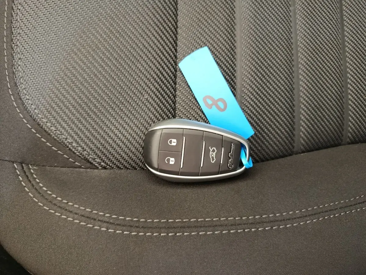 Gros plan sur clé de voiture Alfa Romeo Stelvio posée sur siège tissu gris foncé avec étiquette bleue.