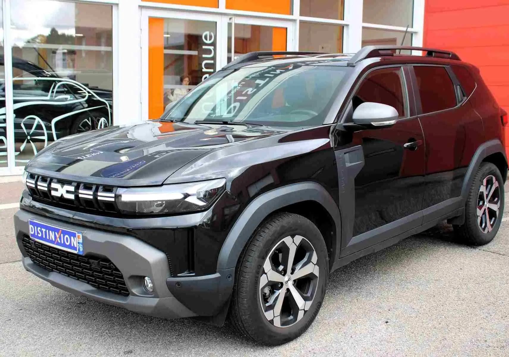 Dacia Duster III noir nacré en 3/4 avant droit, avec jantes alliage et barres de toit noires visibles.