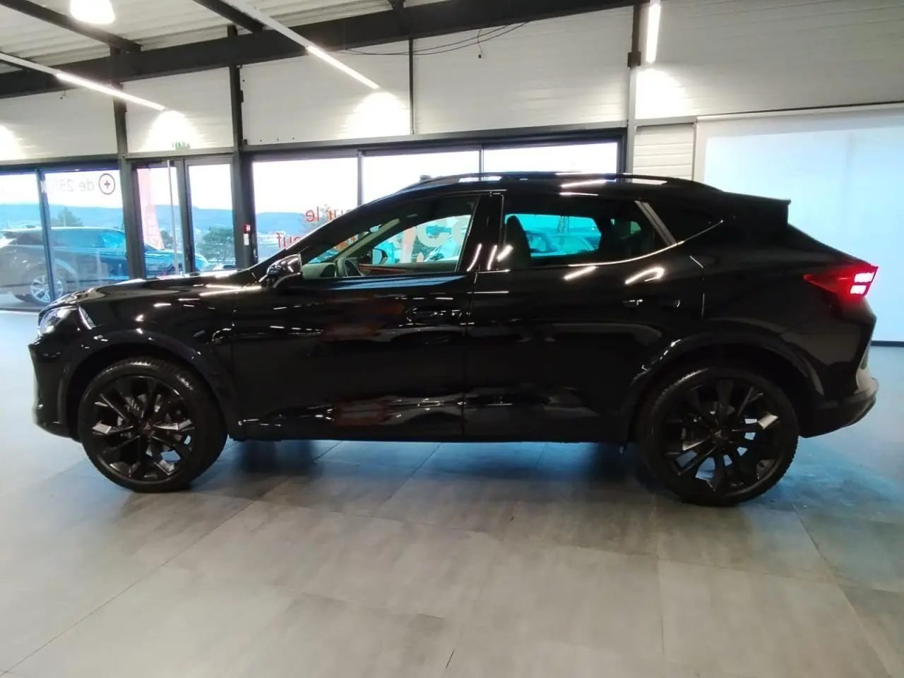 Profil droit du CUPRA Formentor noir métal avec jantes noires et feux arrière allumés en intérieur showroom.