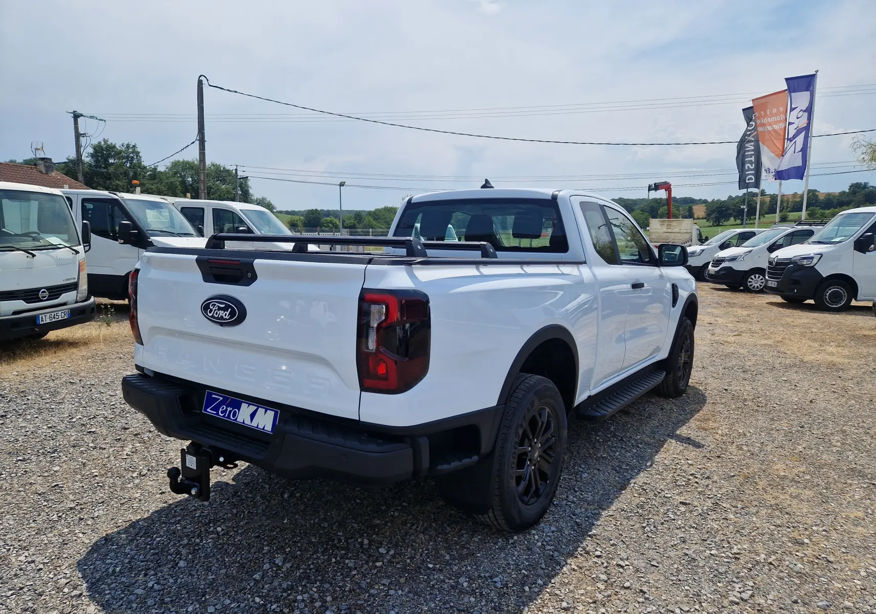Vue 3/4 arrière droite du Ford Ranger Super Cabine blanc avec plateau, barres de toit et attelage visible sur un terrain gravillonné.