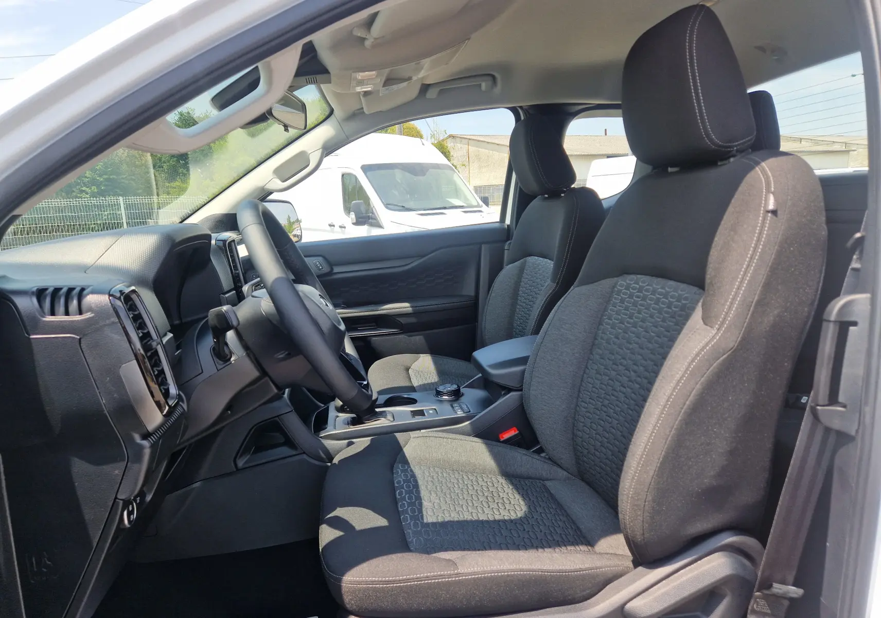 Vue intérieure côté conducteur du Ford Ranger Super Cabine blanc, sièges tissu noir avec motifs et console centrale visible.