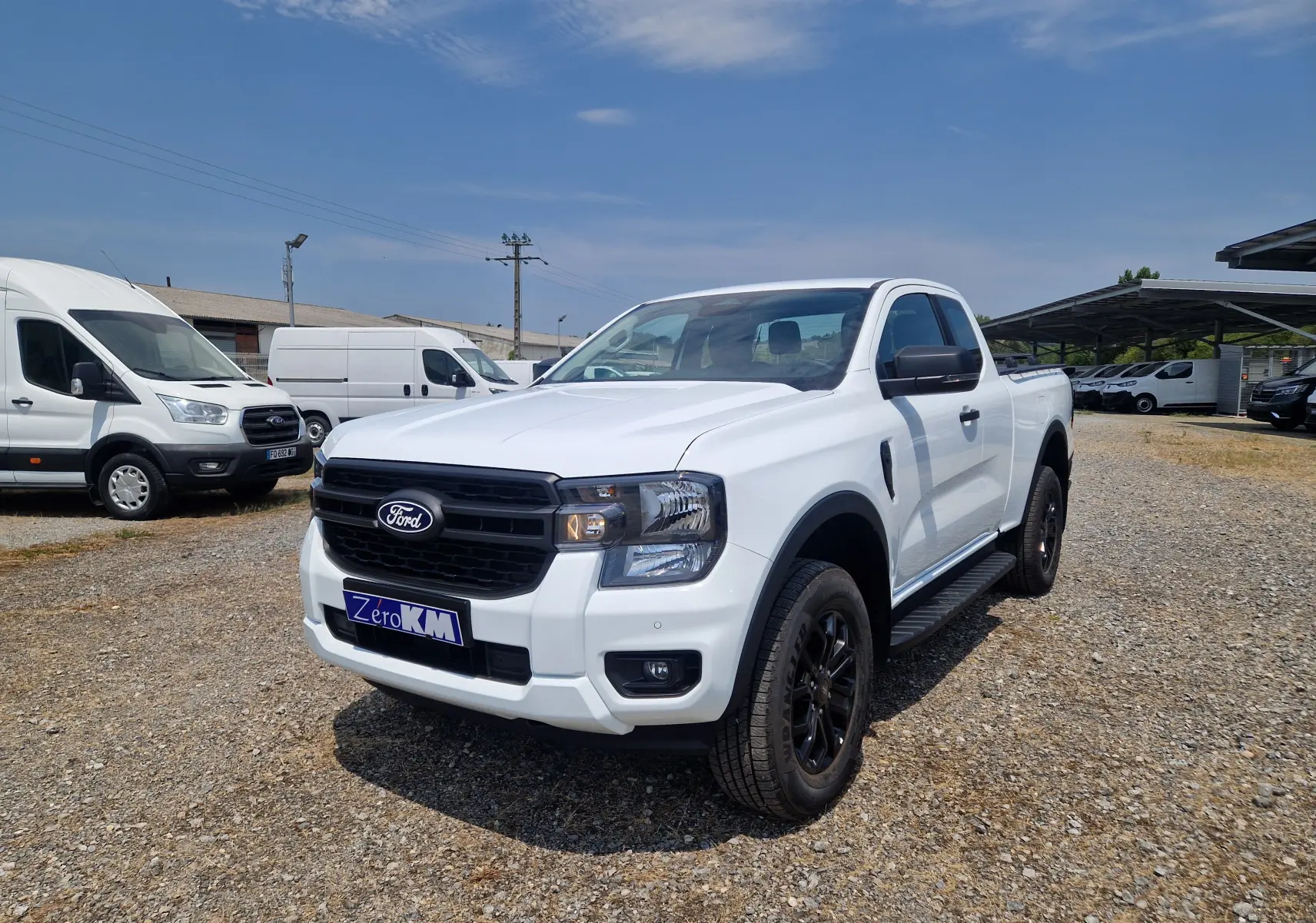 Ford Ranger Super Cabine blanc vu en 3/4 avant droit avec marchepieds noirs et jantes foncées sur terrain gravillonné.