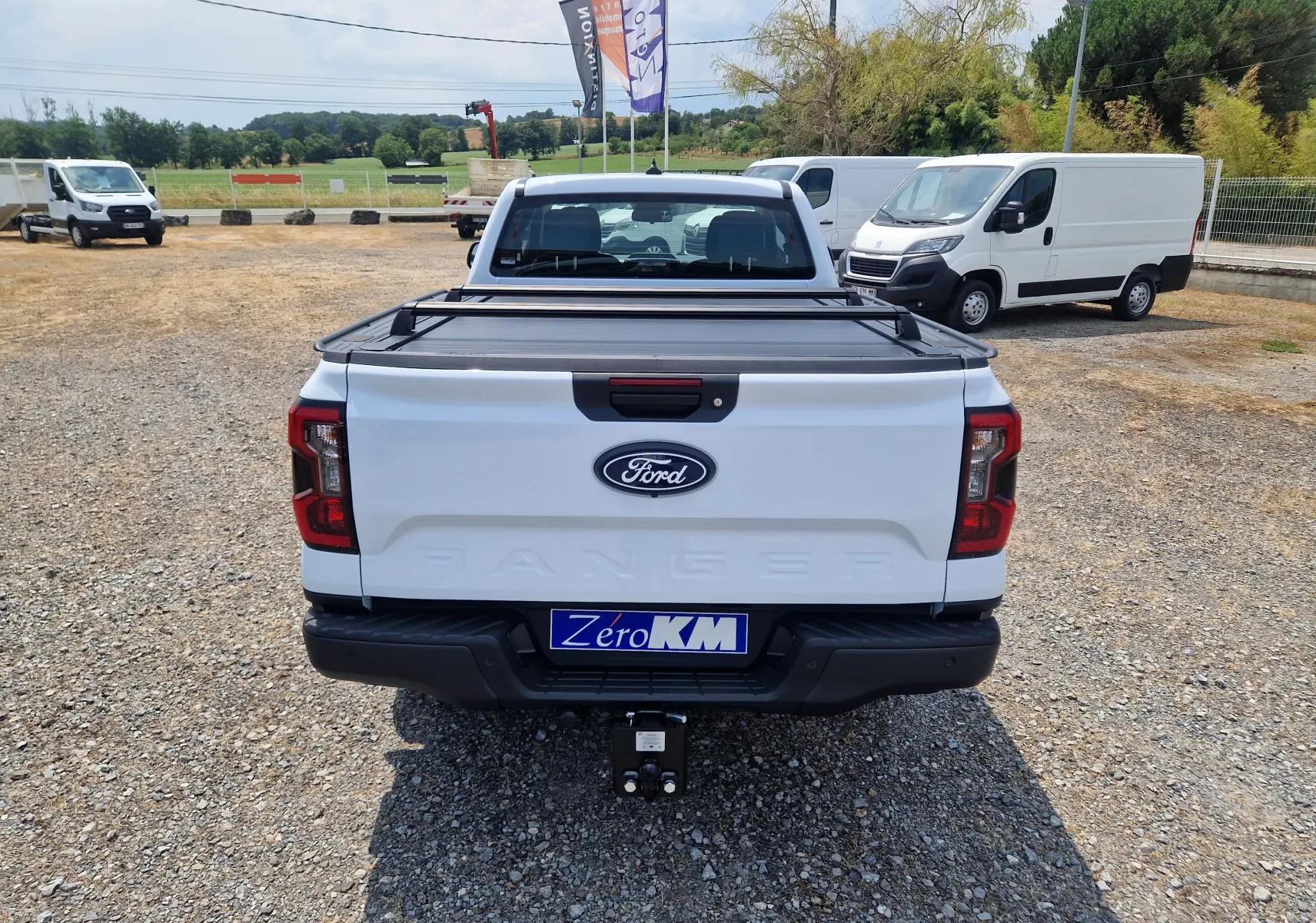 Vue arrière du Ford Ranger Super Cabine blanc avec rideau de plateau rigide et attelage visible sur un terrain gravillonné.
