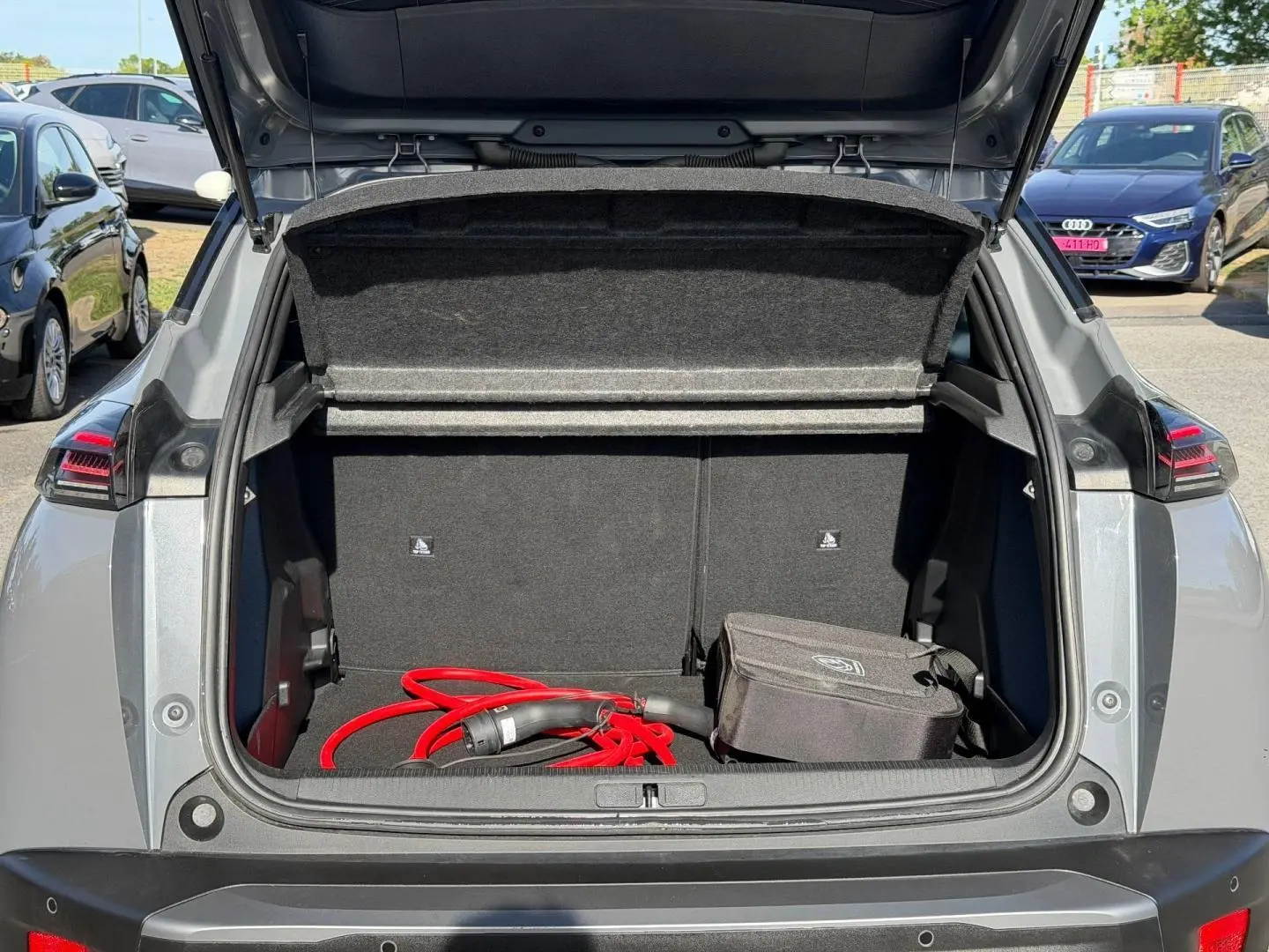 Coffre ouvert en vue arrière d’un Peugeot e-2008 gris Artense avec câble de recharge électrique rouge visible.