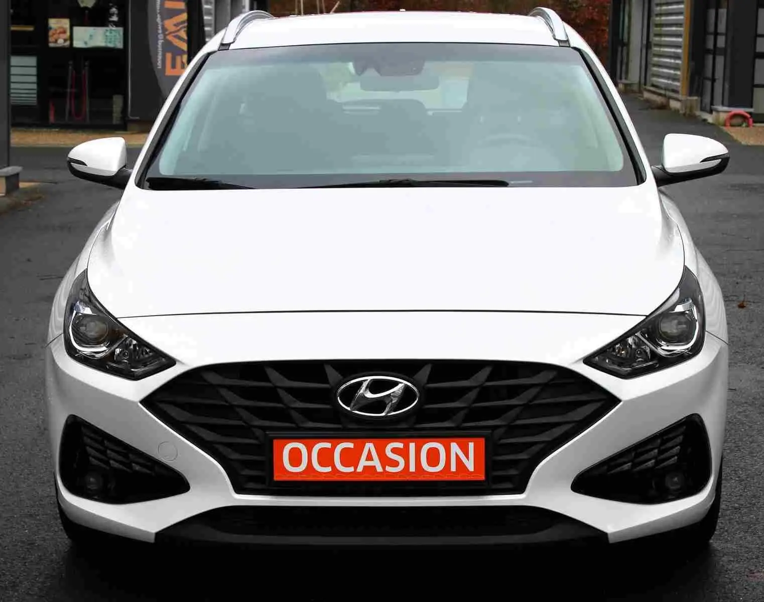 Vue frontale d'une Hyundai i30 SW blanche polaire avec calandre noire et plaque "OCCASION" rouge visible.