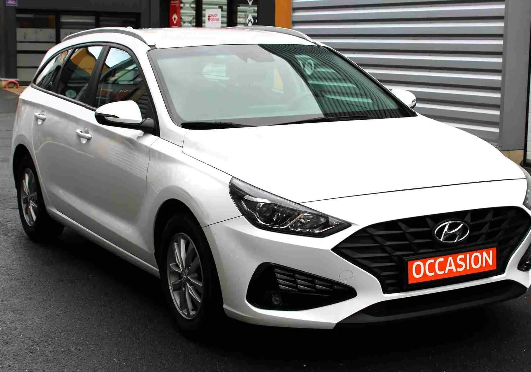 Vue 3/4 avant d'une Hyundai i30 SW blanche polaire avec calandre noire et plaque "OCCASION" rouge visible.