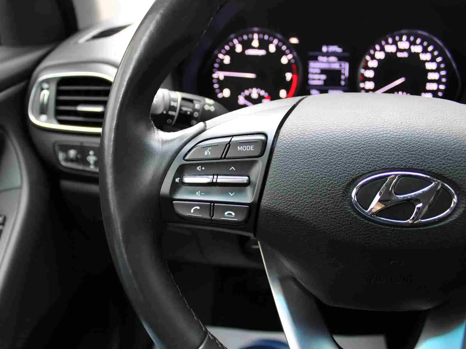 Gros plan sur le volant noir de la Hyundai i30 SW 2022 avec commandes audio et tableau de bord éclairé en arrière-plan.