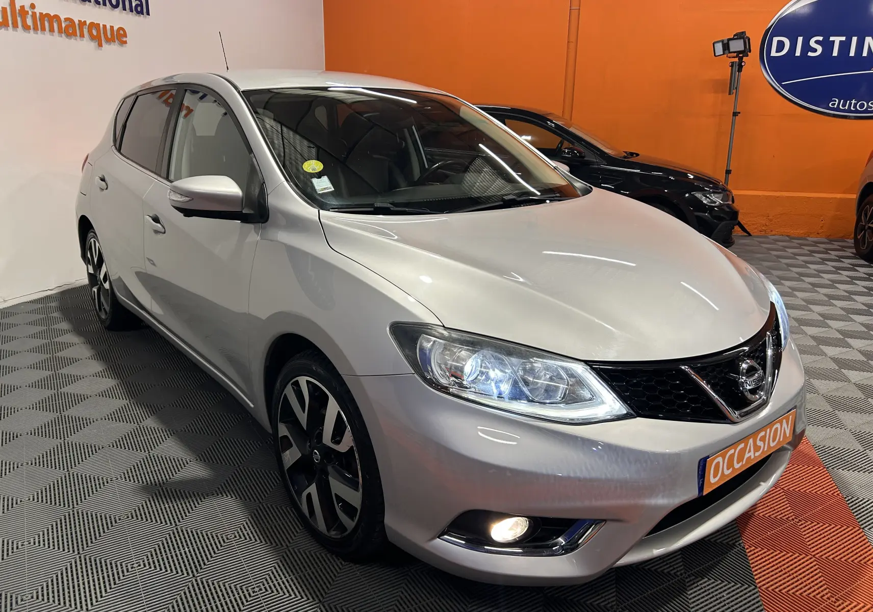 Nissan Pulsar gris vue 3/4 avant droit en intérieur, avec phares allumés et jantes noires et argent.