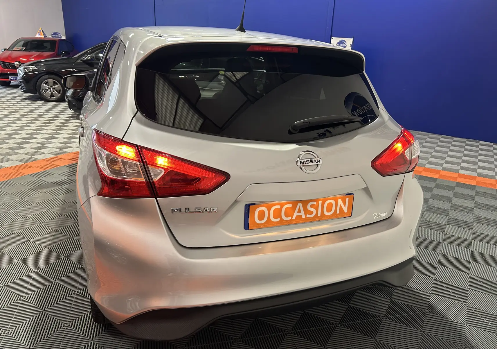 Vue 3/4 arrière d'une Nissan Pulsar gris métallisé avec feux arrière allumés et plaque "OCCASION" en intérieur showroom.