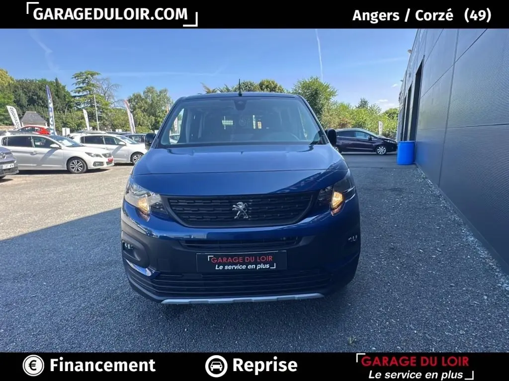 Vue avant d'un Peugeot Rifter bleu 2024 avec calandre noire et feu avant droit allumé sur parking extérieur.