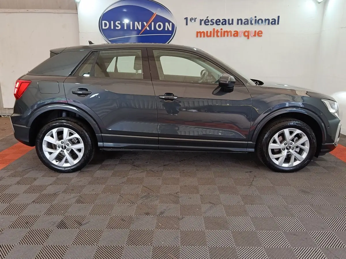 Profil côté gauche d'un Audi Q2 gris foncé 2022, version Business Line, sur sol intérieur avec logo Distinxion en arrière-plan.