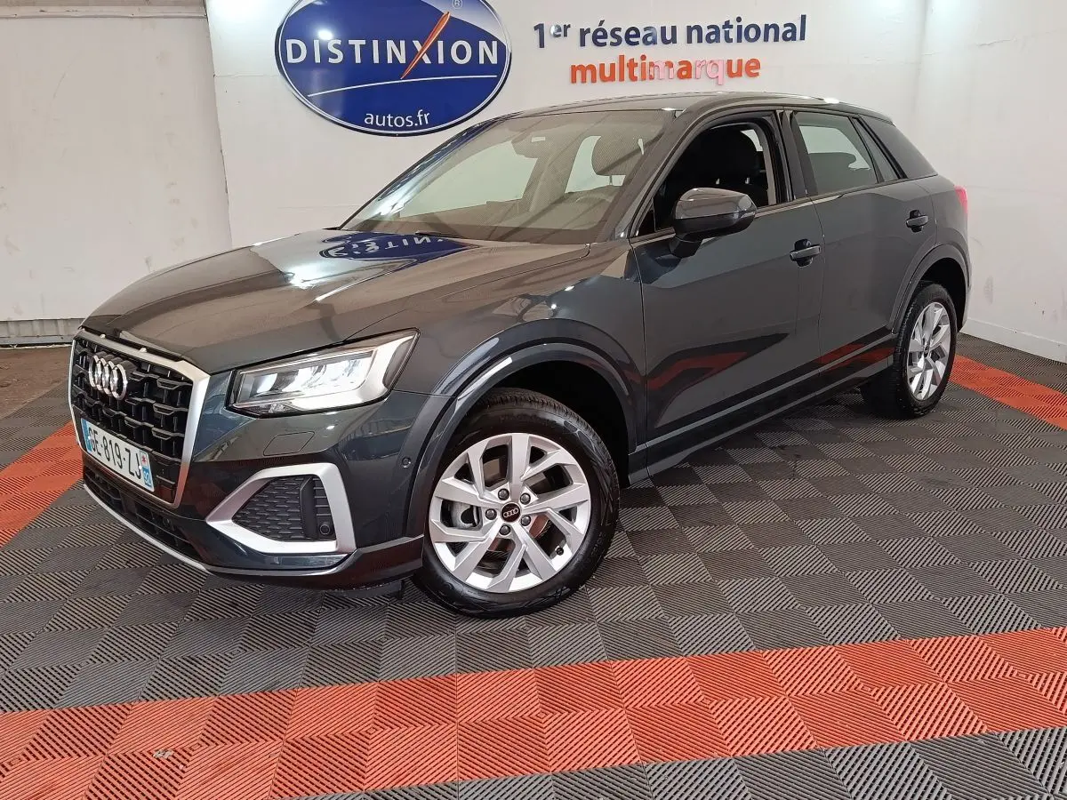 Audi Q2 gris foncé vue 3/4 avant droit, avec jantes alliage et phares LED allumés en intérieur showroom.