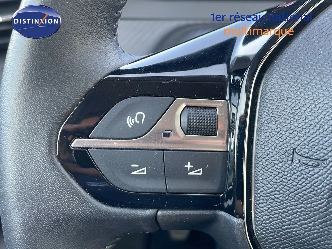 Gros plan sur les commandes de volume au volant noir de la Peugeot 208 blanc banquise, version 2024 essence manuelle.