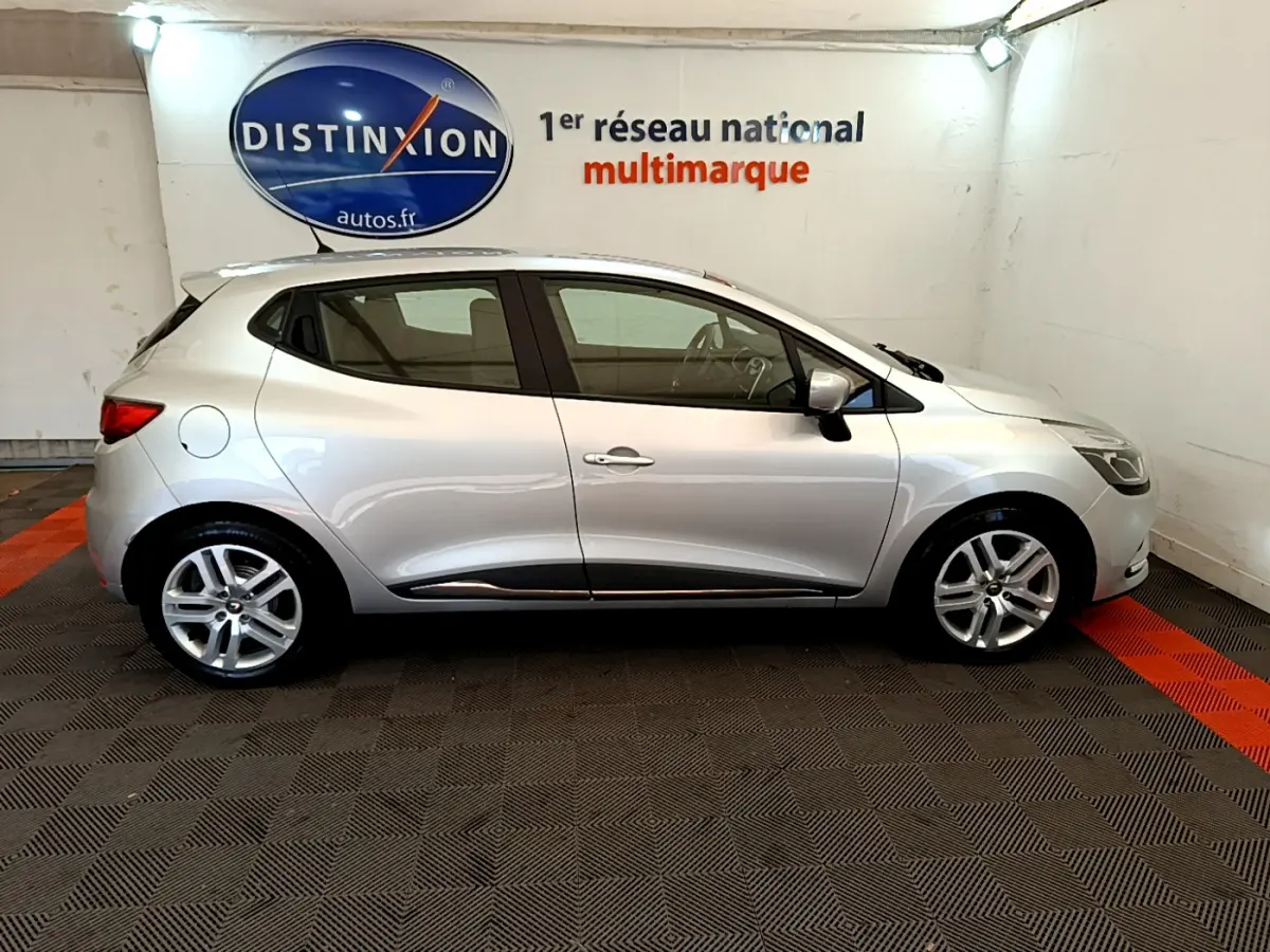 Profil côté droit d'une Renault Clio Business TCe 90 gris clair, en intérieur sur sol damier avec logo Distinxion en arrière-plan.