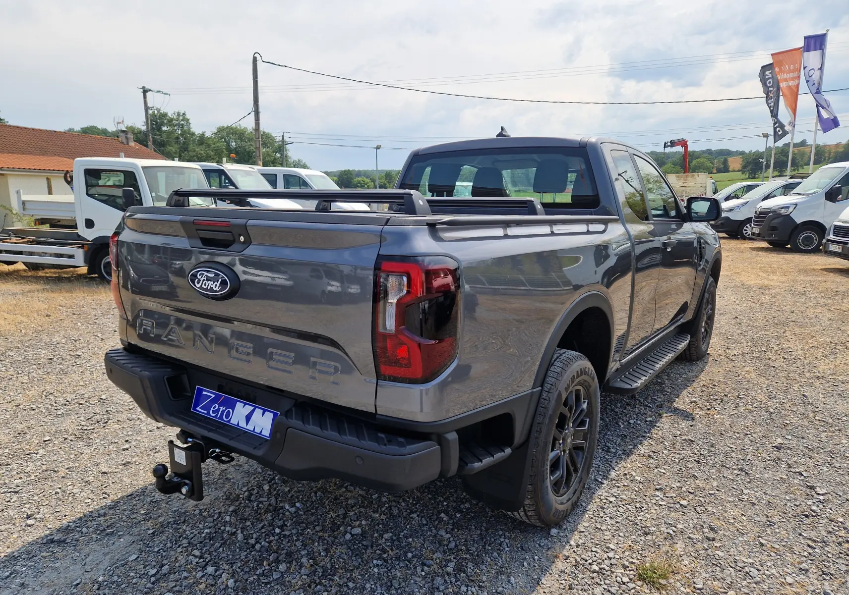 Ford Ranger Super Cabine gris carbone vu en 3/4 arrière droit, avec rideau de plateau rigide et barres de toit visibles.