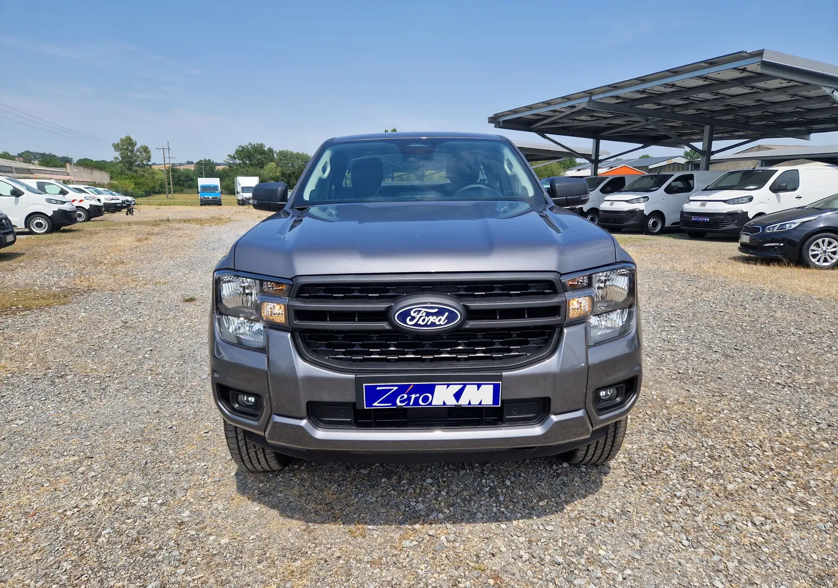 Vue avant d'un Ford Ranger Super Cabine gris carbone avec calandre noire et plaque "Zéro KM" sur un parking extérieur.