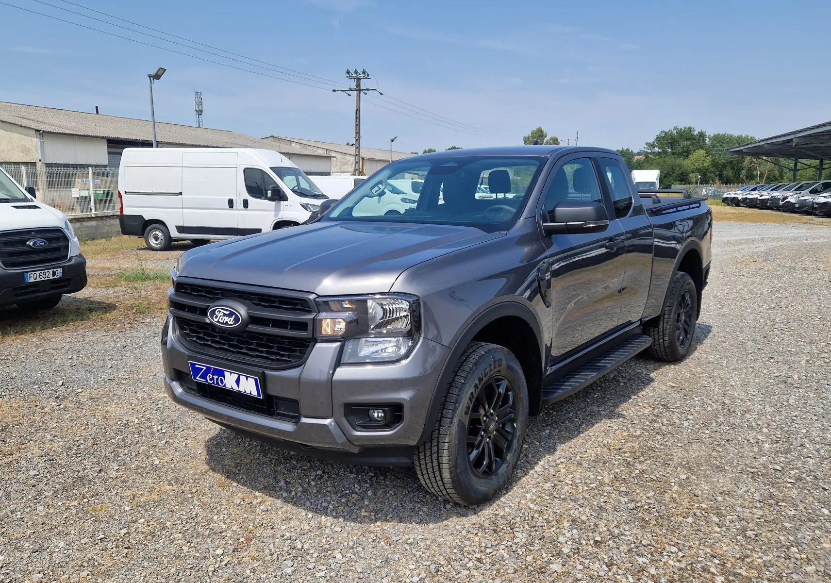 Ford Ranger Super Cabine gris carbone en 3/4 avant droit, avec marchepied noir et jantes noires sur terrain gravelé.