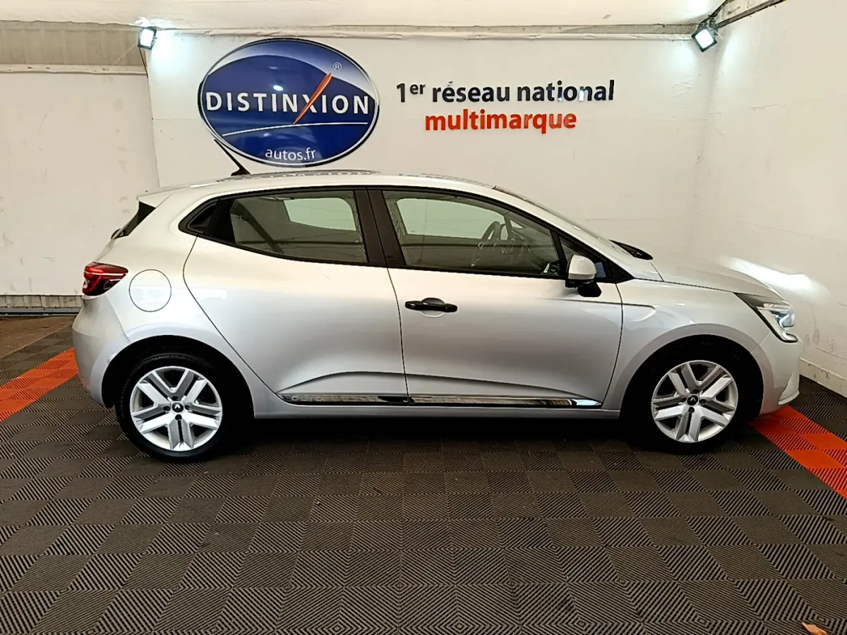Profil droit d'une Renault Clio Business TCe 100 gris clair 2020 dans un showroom avec logo Distinxion en arrière-plan.