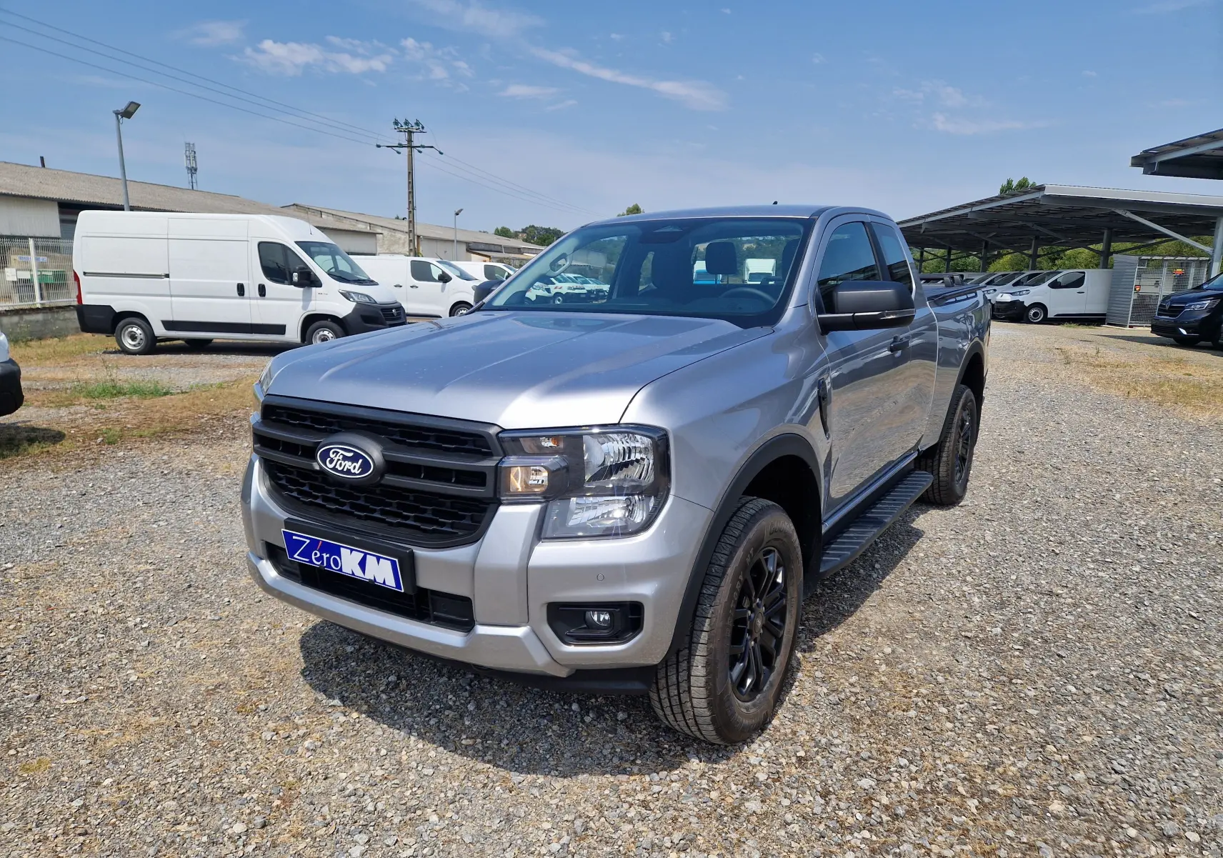 Vue 3/4 avant d'un Ford Ranger Super Cabine gris iconic avec calandre noire et marchepieds sur un terrain gravillonné.