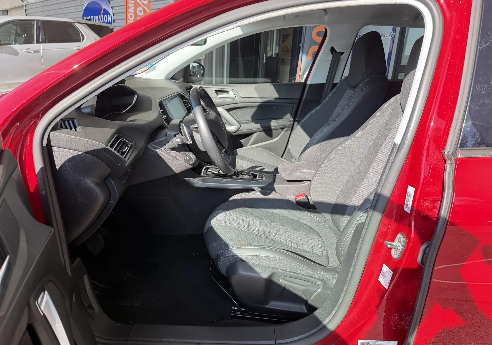 Vue intérieure côté conducteur du Peugeot 308 SW rouge rubis, mettant en valeur les sièges gris et le tableau de bord moderne.