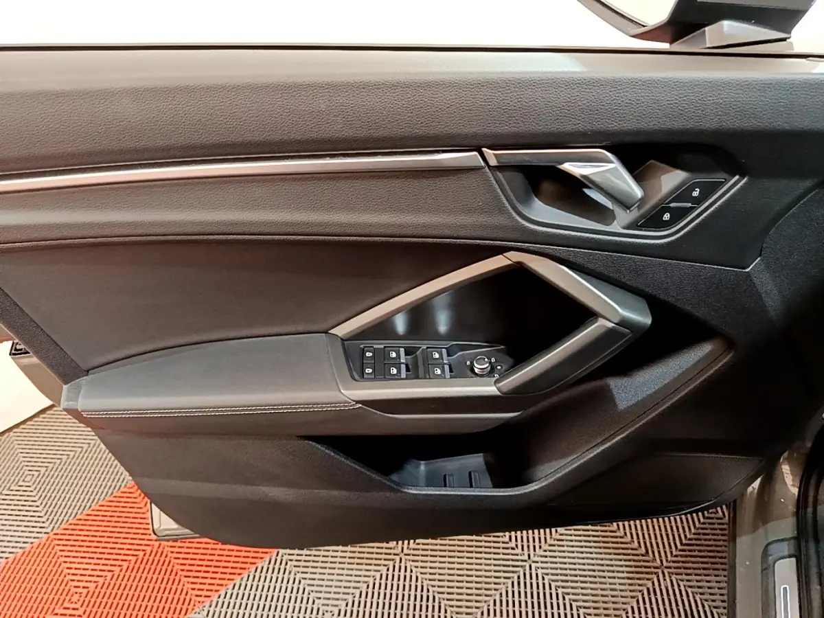 Vue intérieure côté gauche de la porte avant d’un Audi Q3 gris foncé avec commandes de vitres et poignée argentée.