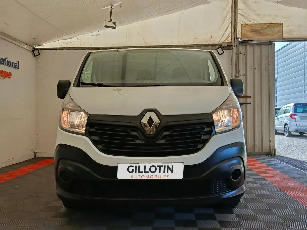 Vue frontale d'un Renault Trafic Fourgon blanc avec calandre noire et phares allumés dans un garage.