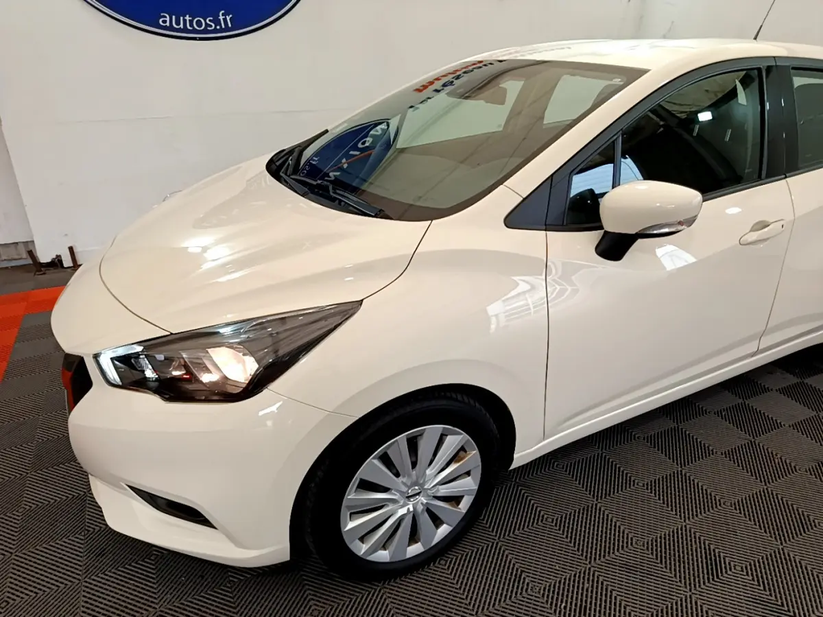 Vue 3/4 avant droit d'une Nissan Micra IG-T 92 Acenta blanche avec phares allumés et rétroviseurs intégrant clignotants.