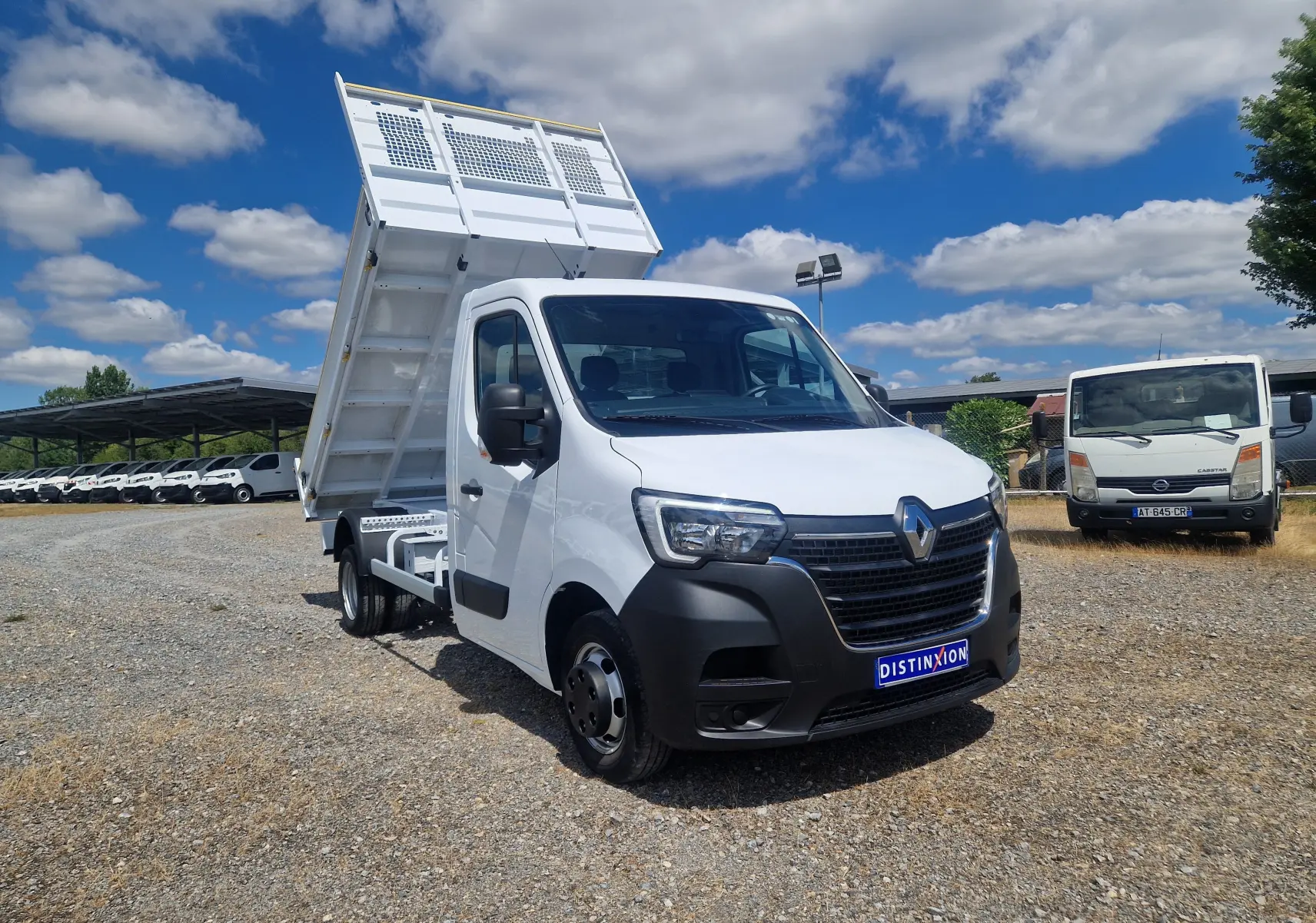 Renault Master Benne blanc en 3/4 avant droit, benne relevée et roues jumelées visibles sur terrain gravillonné.