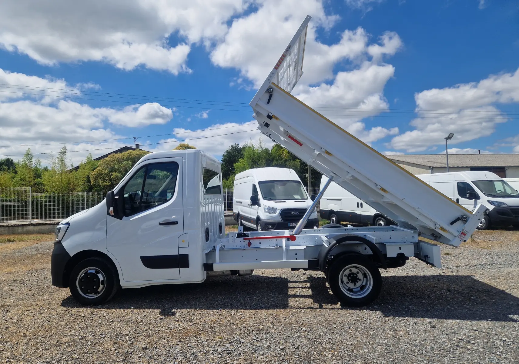 Vue latérale gauche du Renault Master Châssis Cabine blanc 2024 avec benne relevée et roues jumelées sur terrain gravelé.