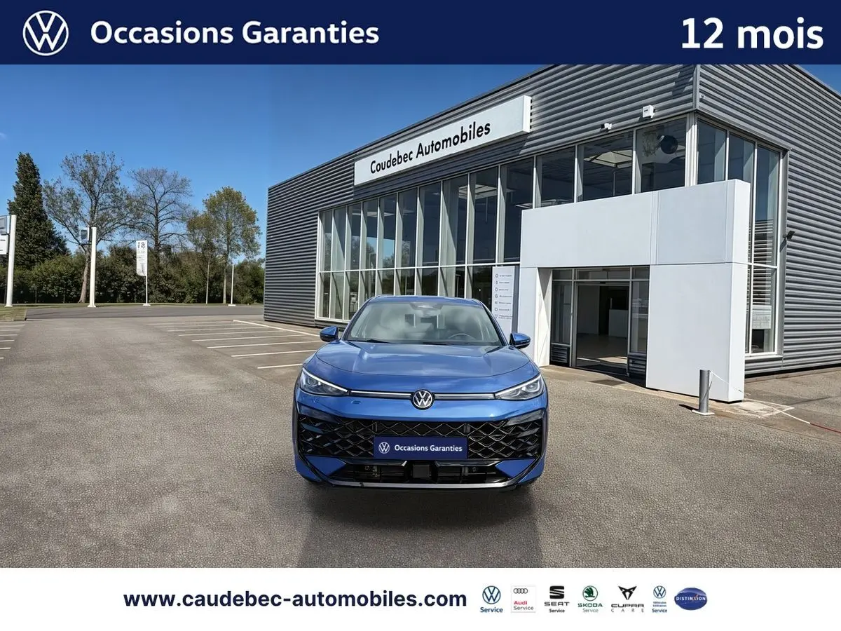 Volkswagen T-Roc bleu métallisé vu de face, avec calandre noire et logo lumineux devant un bâtiment moderne.