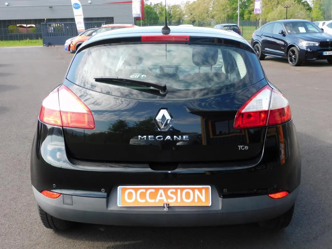 Vue arrière d'une Renault Mégane noire de 2015 avec logo et plaque "OCCASION" visible sur un parking.