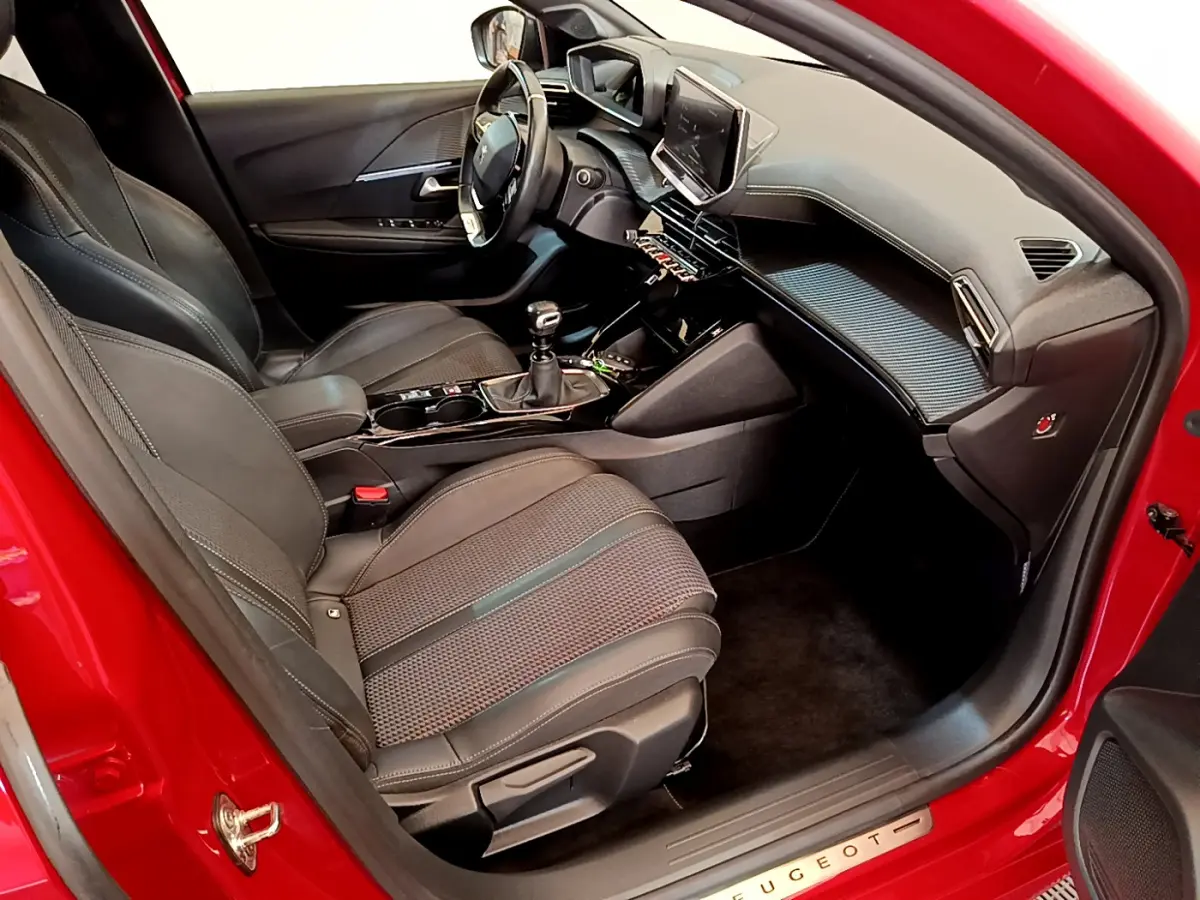 Intérieur côté conducteur d'une Peugeot 208 rouge, montrant sièges noirs, volant cuir et écran tactile central.