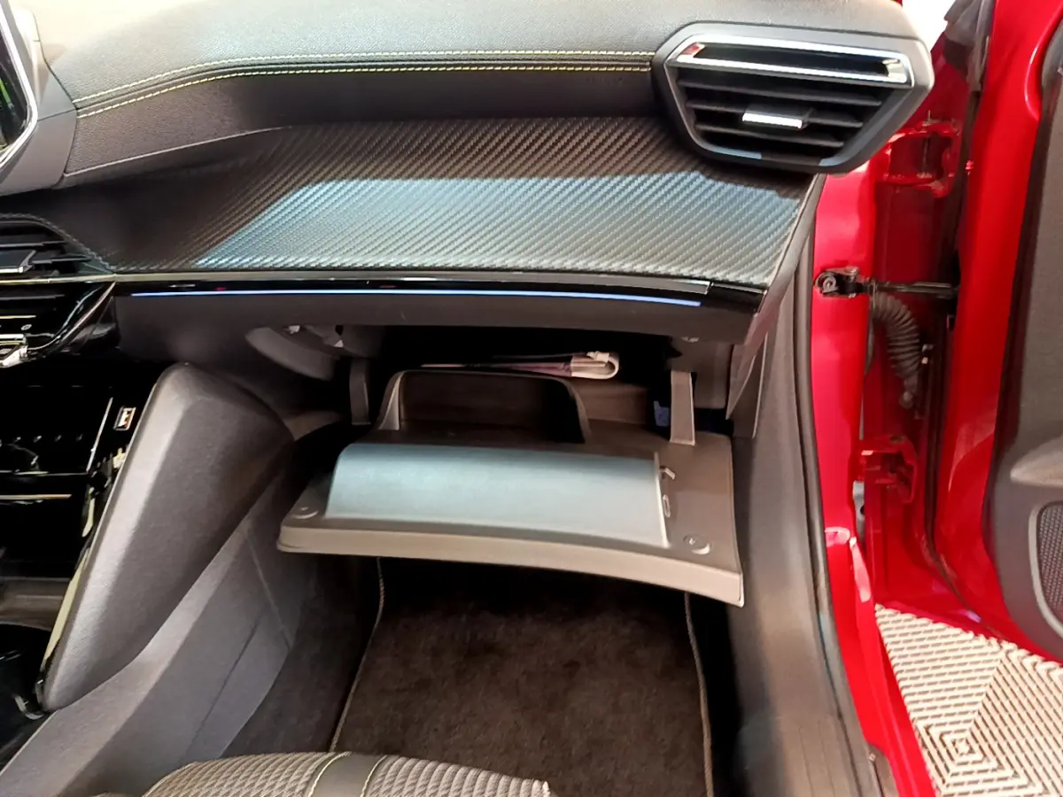 Vue intérieure côté passager d'une Peugeot 208 rouge 2020 avec boîte à gants ouverte et tableau de bord noir à insert carbone.