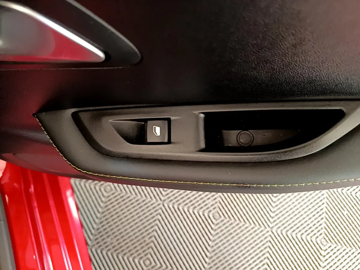 Gros plan sur la commande de lève-vitre côté gauche dans la porte intérieure noire d'une Peugeot 208 rouge.