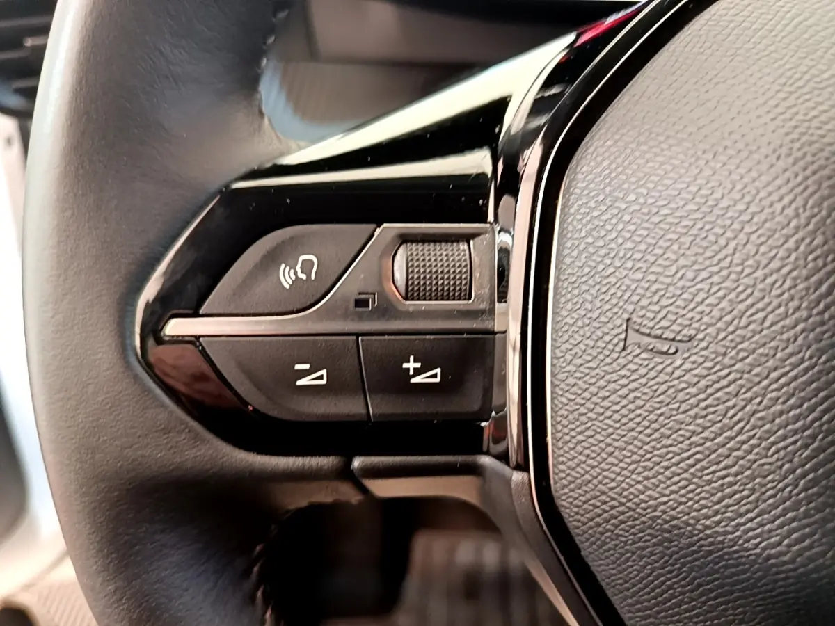 Gros plan sur les commandes gauche du volant cuir noir du Peugeot 208 blanc, avec boutons multifonctions.