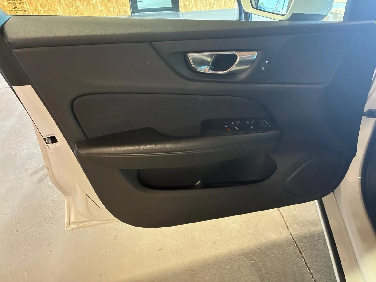 Vue intérieure de la porte avant gauche noire d’une Volvo V60 blanche, avec commandes de vitres et poignée métal.