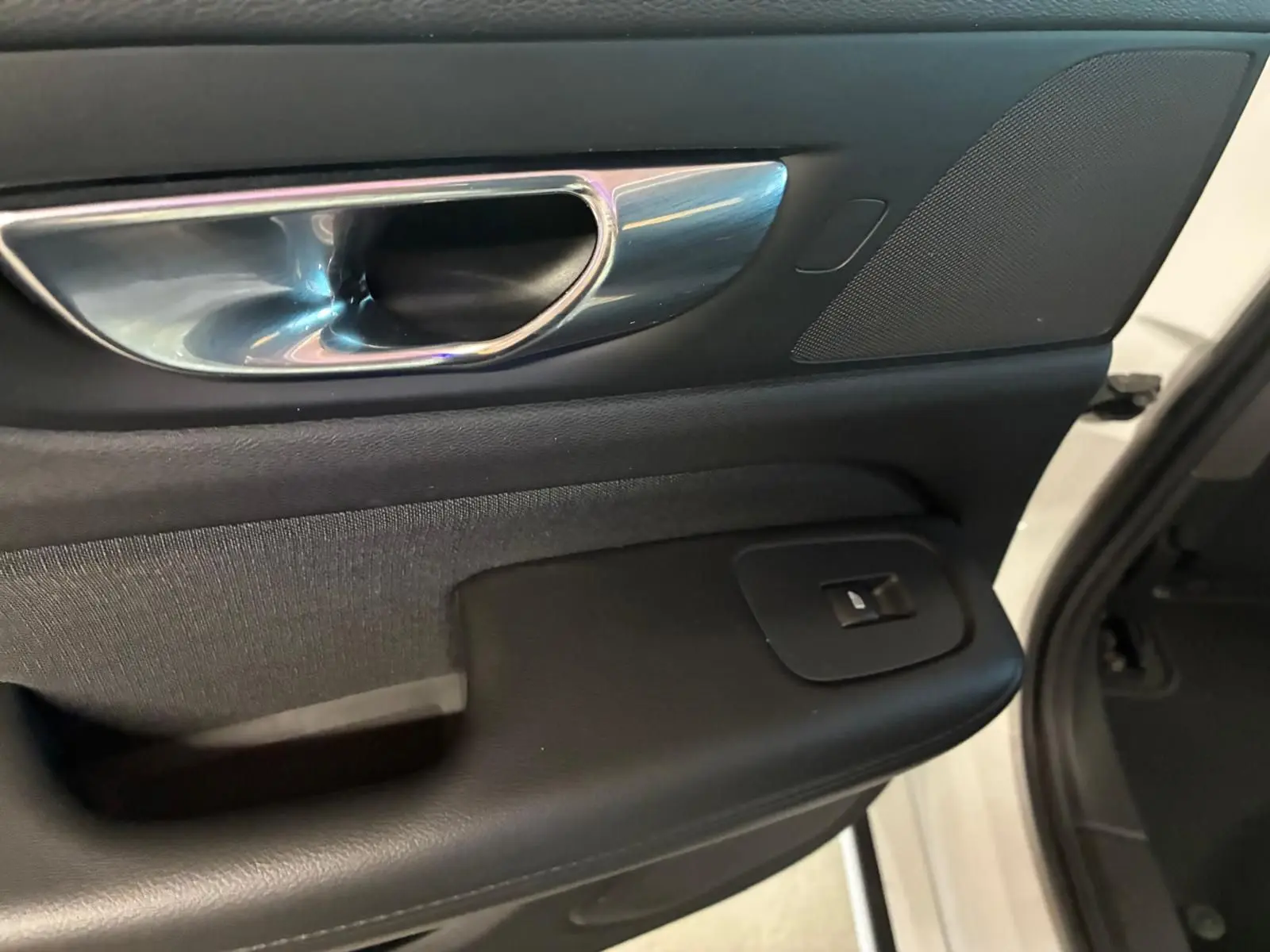 Poignée intérieure côté gauche de la porte avant d'une Volvo V60 blanche, avec bouton de commande de vitre et finition noire.
