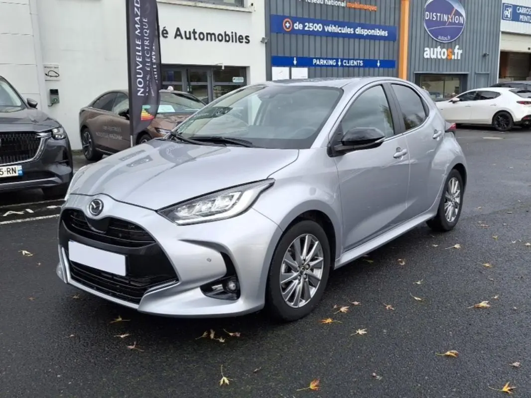Vue 3/4 avant d'une Mazda2 Hybrid argent 2022 garée devant un concessionnaire, avec calandre noire et phares LED.