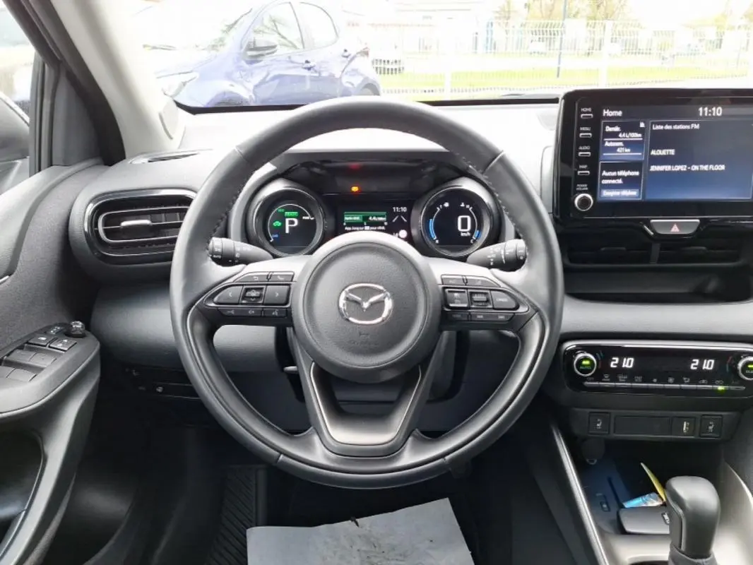 Vue intérieure centrée sur le volant de la Mazda2 hybride 2022, avec tableau de bord numérique et écran tactile visible.