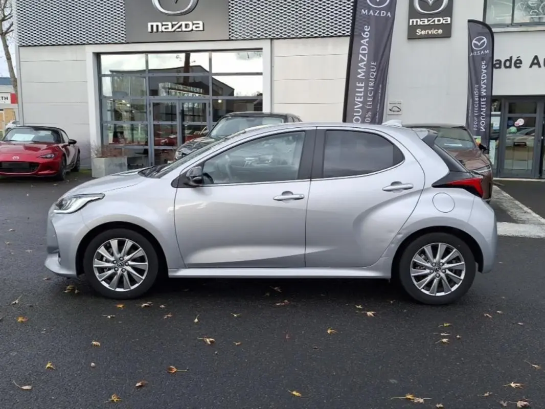 Profil côté gauche d'une Mazda2 hybride 2022 argent garée devant un concessionnaire Mazda avec jantes alliage.