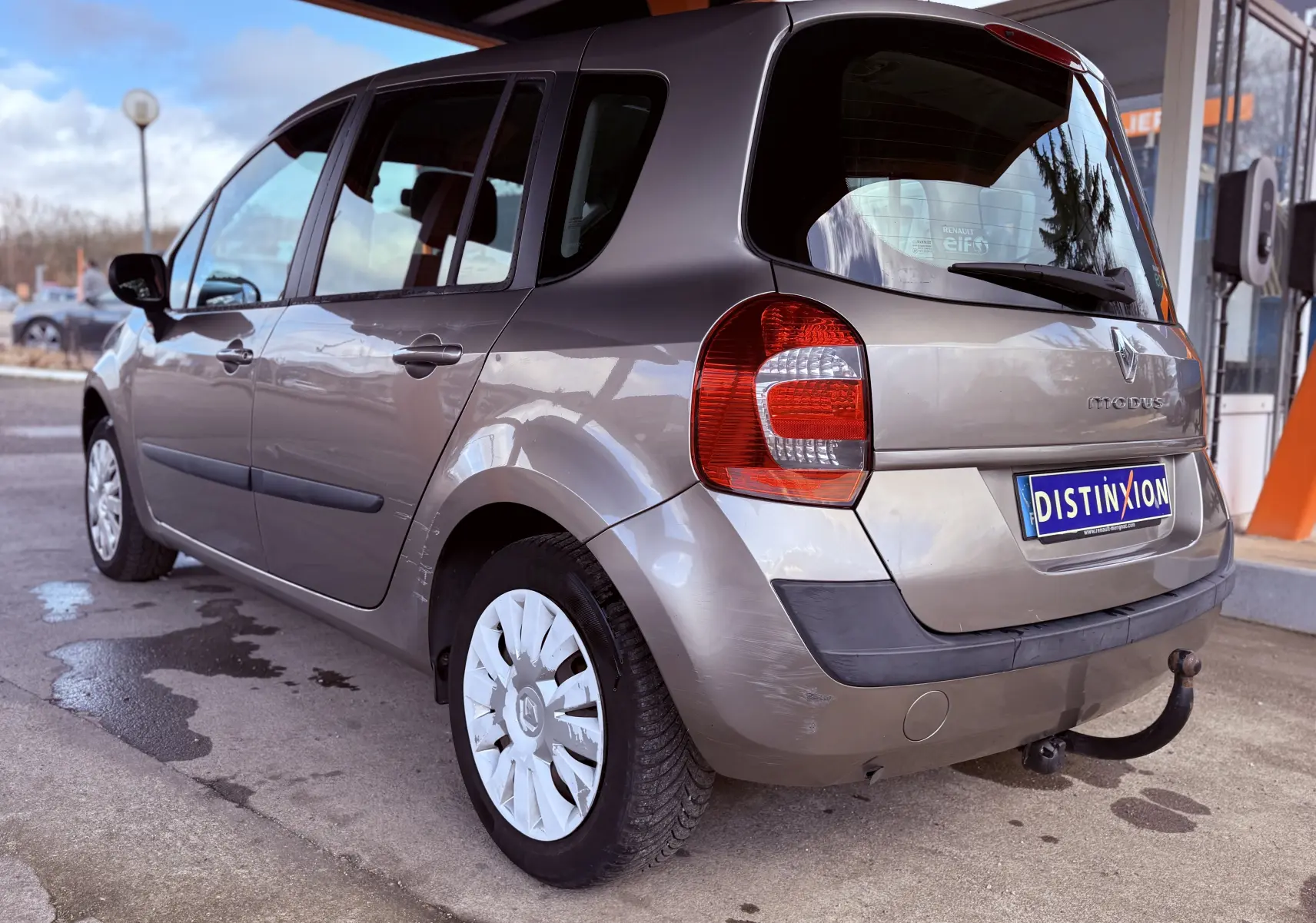 Vue 3/4 arrière gauche d'une Renault Modus beige de 2011 avec attelage et jantes à enjoliveurs blancs.