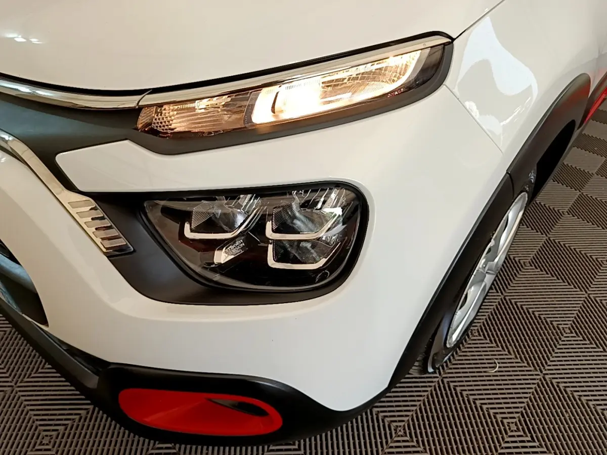 Gros plan sur le phare avant droit allumé d'une Citroën C3 blanche, avec détail rouge sur le pare-chocs noir.