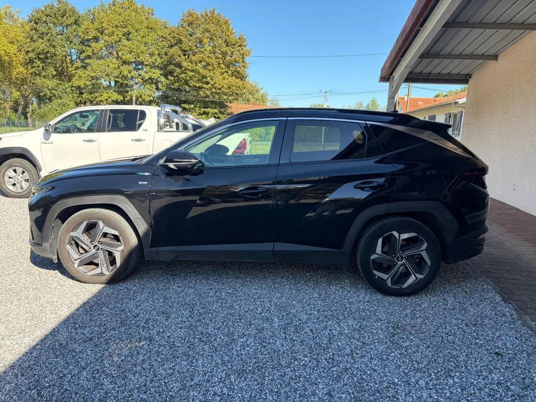 Vue de profil côté gauche d’un Hyundai Tucson noir 2022 garé sur gravier, avec barres de toit et jantes distinctives.