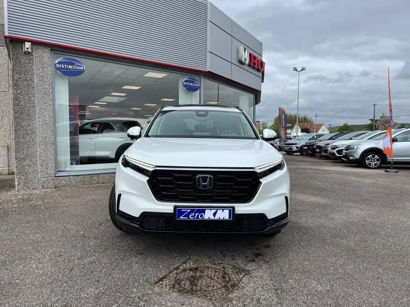 Vue frontale d'un Honda CR-V 2024 blanc platine nacré avec calandre noire et plaque Zero KM devant un concessionnaire.