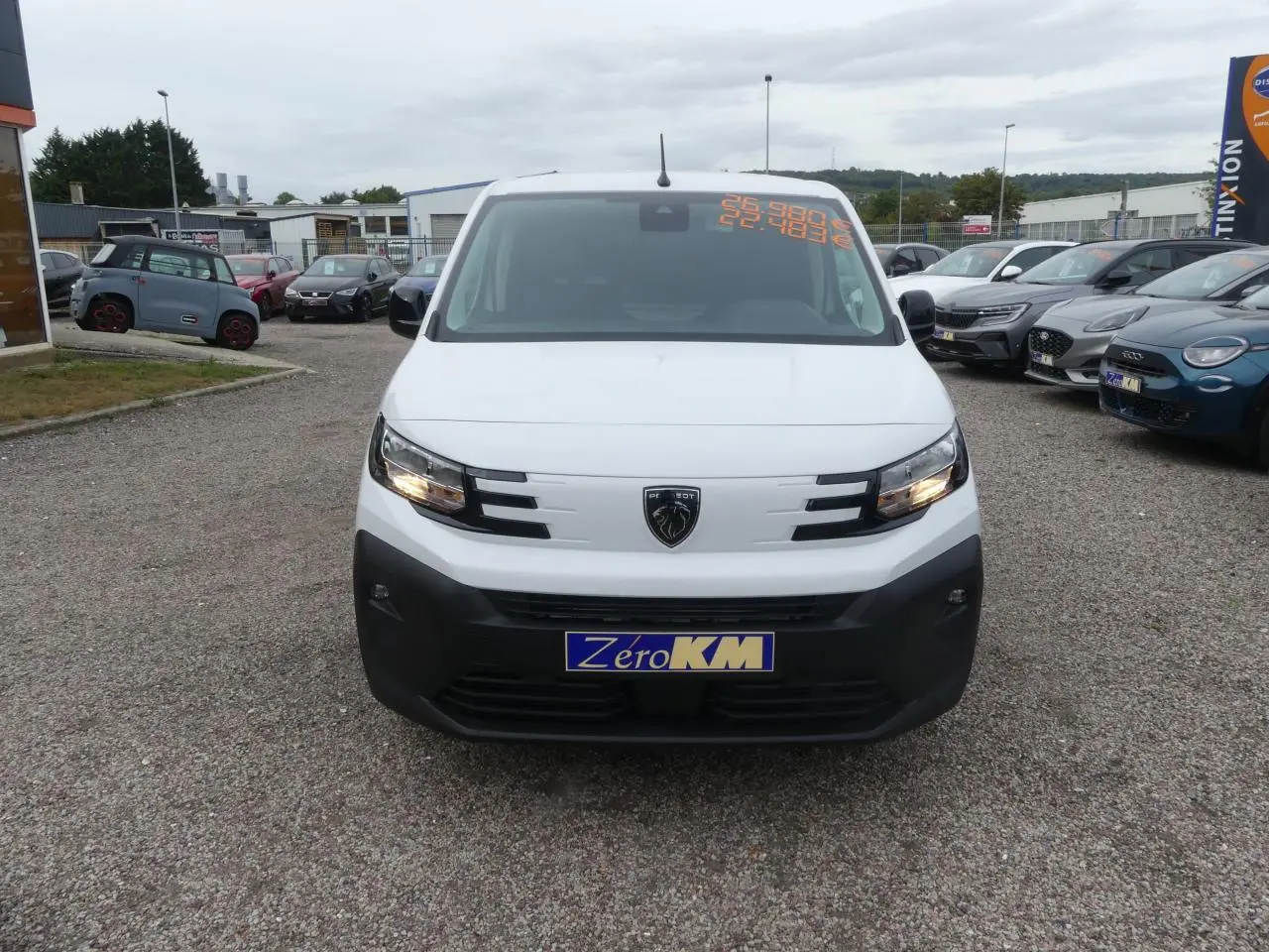 Vue de face d'un Peugeot Partner blanc avec pare-chocs noir et plaque d'immatriculation Zéro KM sur un parking.